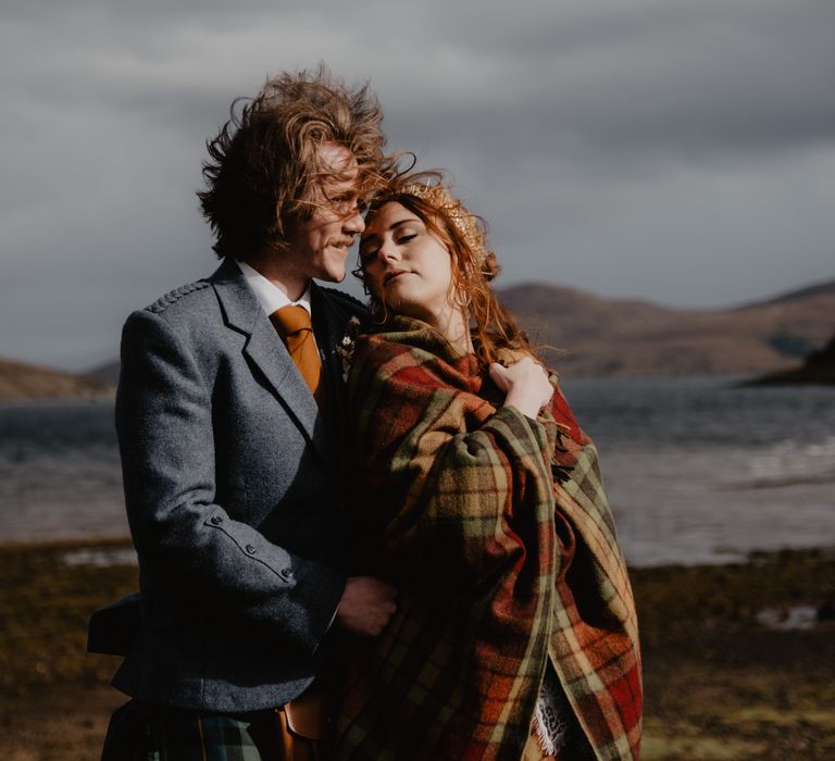 Bride leans back to kiss groom as she wraps tartan blanket around her shoulders