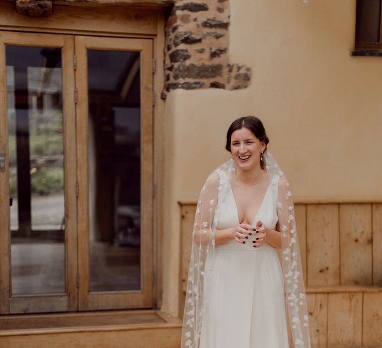 Smiling bride in V neck Charlie Brear wedding dress and daisy chapel length applique veil stands inside before wedding at Hayne, Devon