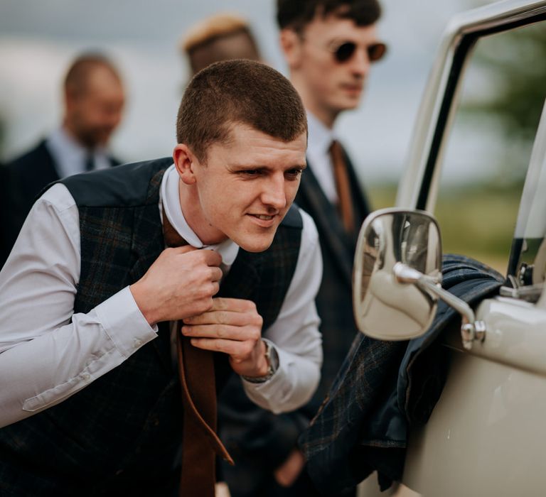 Groom in a navy check waistcoat putting on his brown tie in a car side mirror