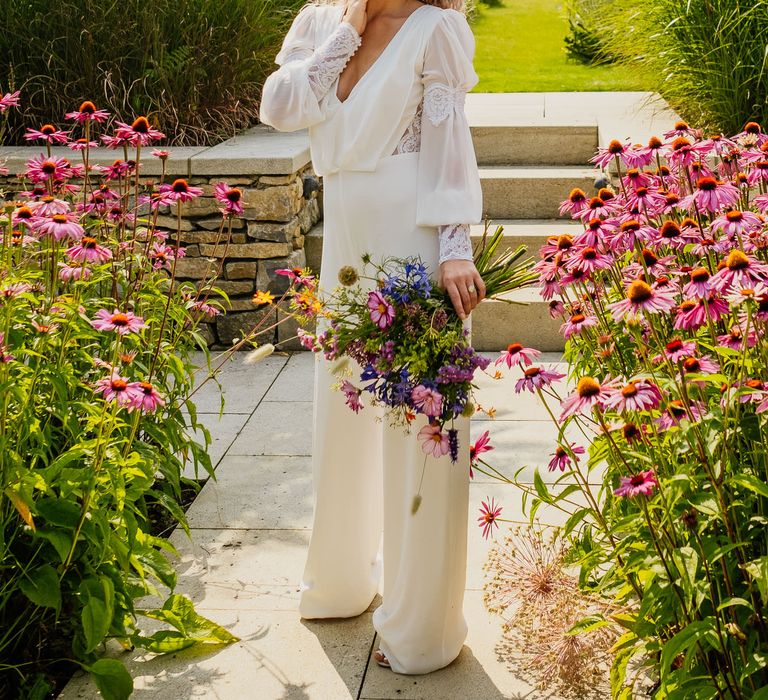 Blonde naturally curly haired bride with bright red lipstick in a jumpsuit with lace cuff sleeve detail holding a wildflower bouquet 