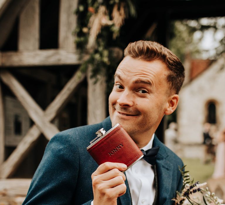 Groom in blue suit and bow tie smiles at the camera whilst holding up red leather personalised hip flask after church wedding ceremony