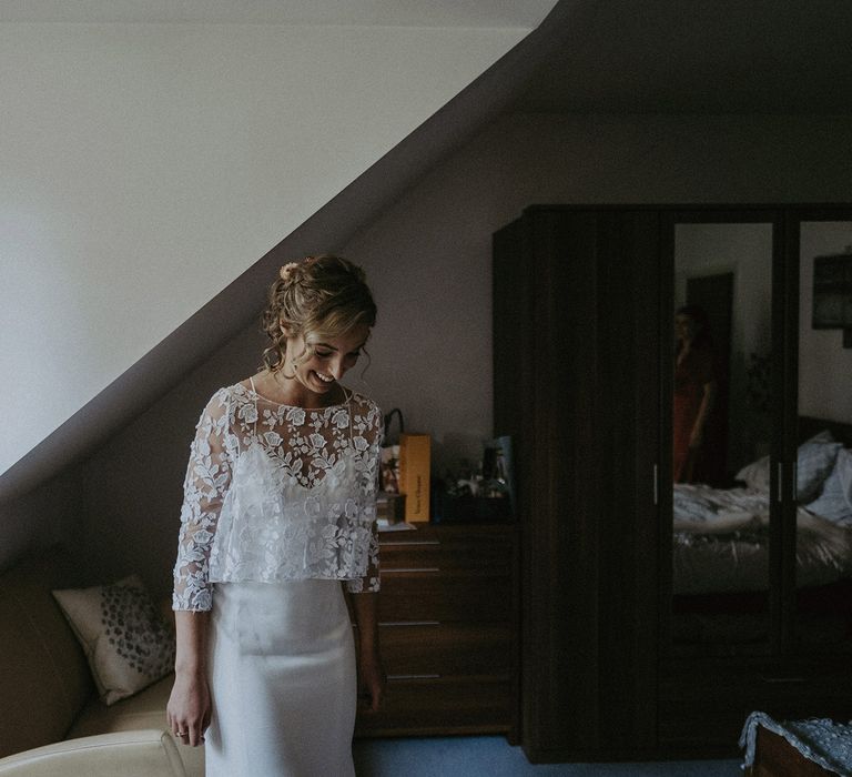 Smiling bride with curled updo in Rime Arodaky wedding dress and mesh embroidered three-quarter sleeve top stands in bedroom before garden wedding with burnt orange wedding theme