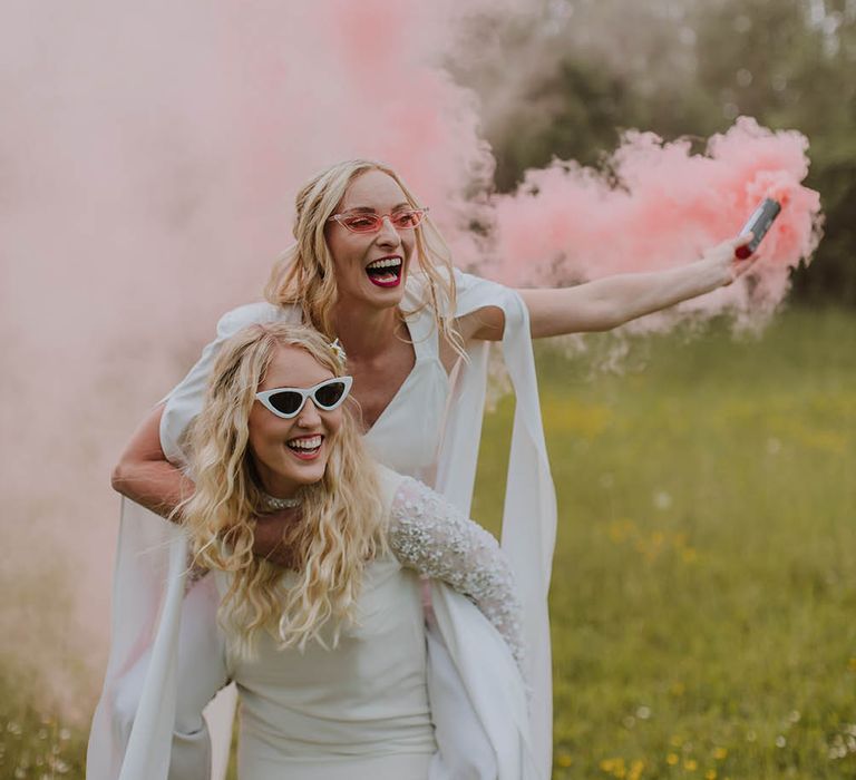 Bride in a short wedding dress giving her bride in a jumpsuit a piggy back as she holds a pink smoke bomb 