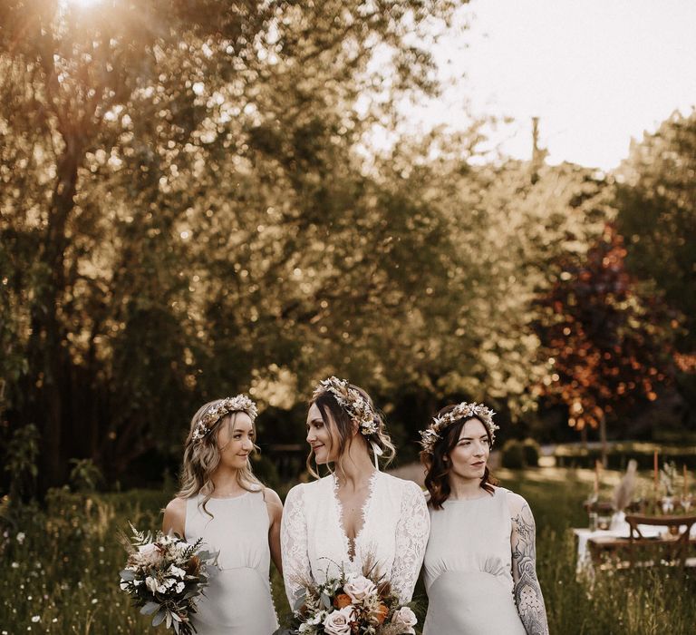 Bride in a long sleeve lace wedding dress with plunging neckline holding a white, green and orange wedding bouquet flanked by her bridesmaids in grey satin dresses 