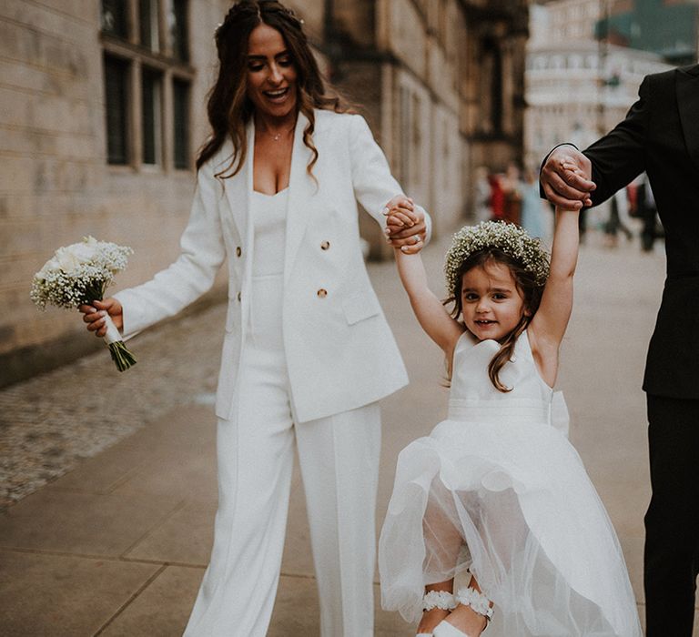 Flower girl in a tulle skirt dress and gypsophila flower crown being swung by her bride and groom parents in Reiss suits 