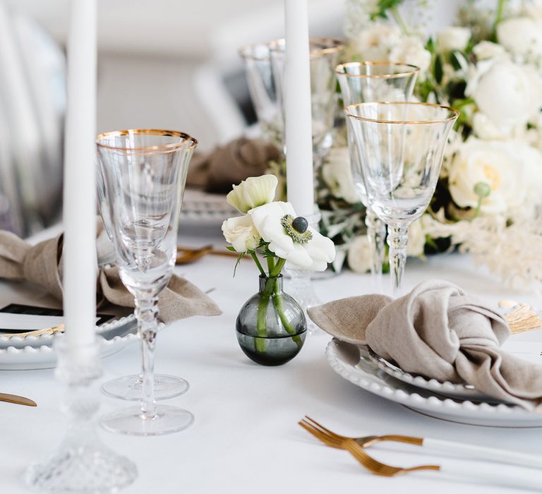 A bud vase filled with lithianthus and anemones sits between gold rimmed glassware and tall white tapered candles at a black and white wedding.. 