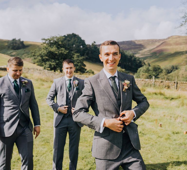 Groom in a mid-grey wedding suit doing up his jacket button with his groomsmen in the background 