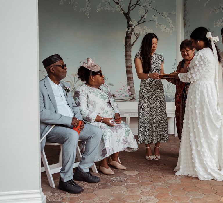 Bride in long mesh sleeve spotted Vagabond wedding dress with bridal hair bow stands next to groom in double breasted linen suit with wedding guests in Victorian summerhouse for traditional Chinese wedding ceremony at Wasing Park wedding