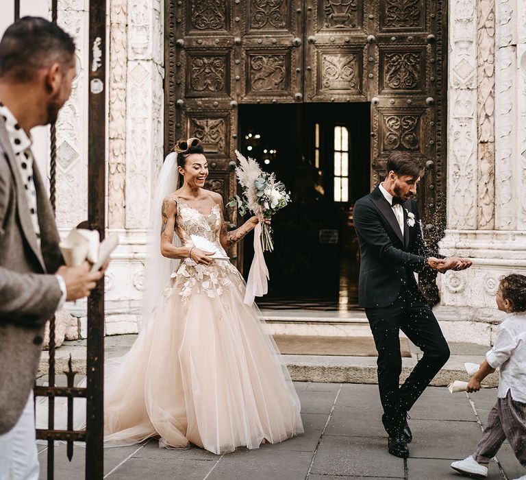 Bride & groom laugh as they throw confetti around their child in front of church on their wedding day