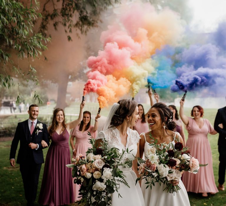 Wedding photo of two brides holding wildflower bouquets with their wedding party following them carrying rainbow coloured smoke flares 