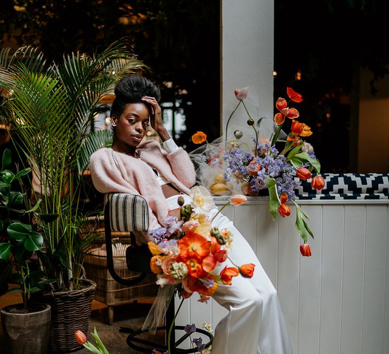 Black bride in trousers and a crop top with a light pink cardigan surrounded by colourful Spring wedding flowers 