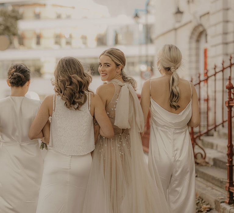 Bride in a halterneck off white wedding dress with bow detail and bridesmaids in white satin wrap dress and jumpsuit walking through London