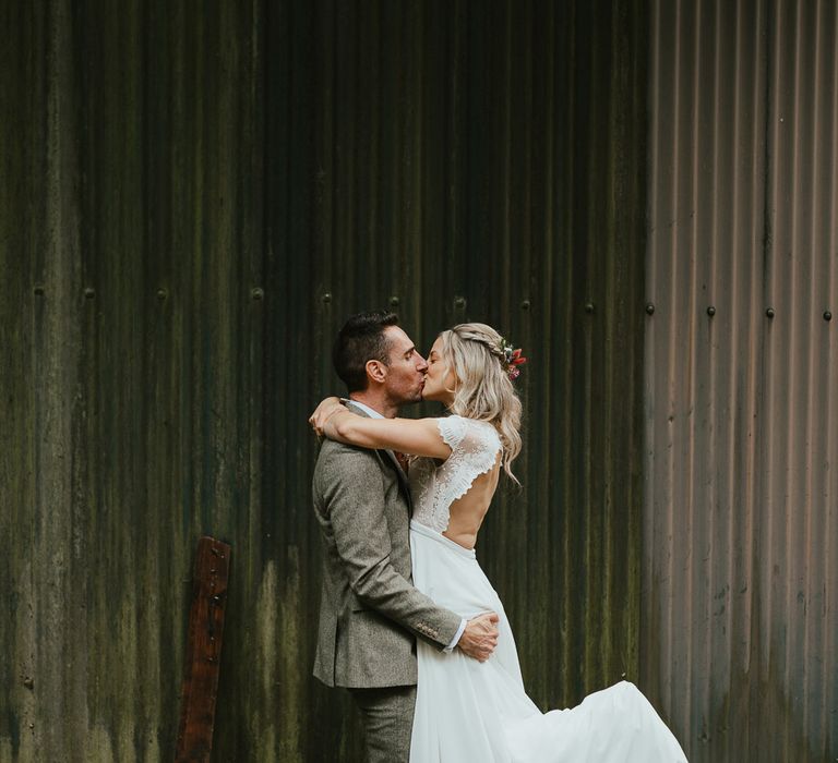 Bride in white lace open back wedding dress kisses groom in grey herringbone suit stood outside at late summer wedding in Norfolk