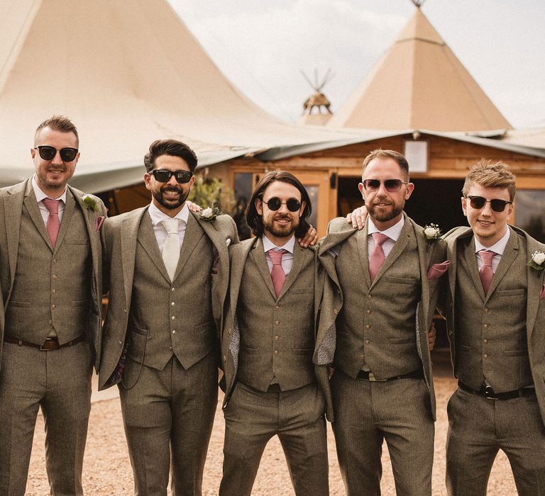 Groom in grey three piece suit with white tie stands with groomsmen in grey three piece suits, pink ties and pink pocket squares all in sunglasses outside tipi at Inkersall Grange Farm