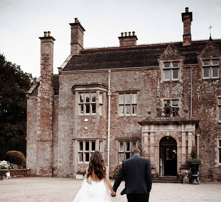 Bride wears Eeysee wedding dress & holds hand with groom as they walk into Cefn Tilla Court 