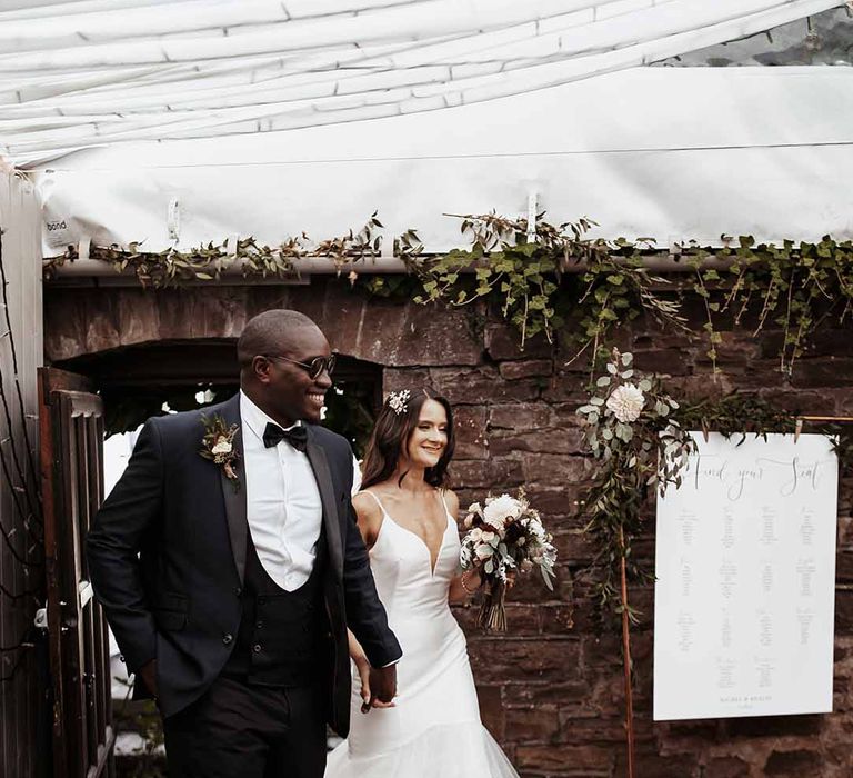 Groom with sunglasses on & three piece suit walks hand in hand with his newly wife to celebrate with guests with wedding signs and trailing ivy in the background 