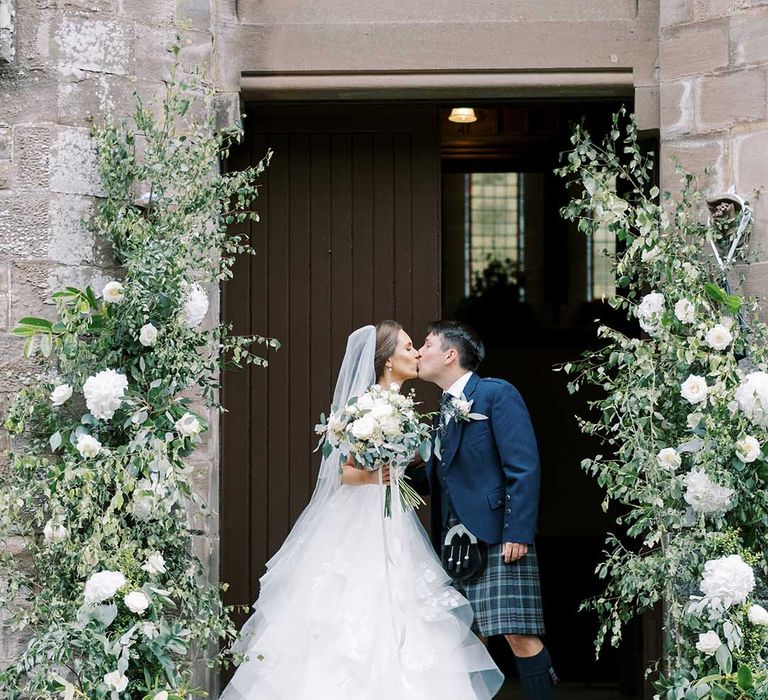 Bride in a Martina Liana princess wedding dress and groom in a wool suit and tart kilt kissing outside the church 