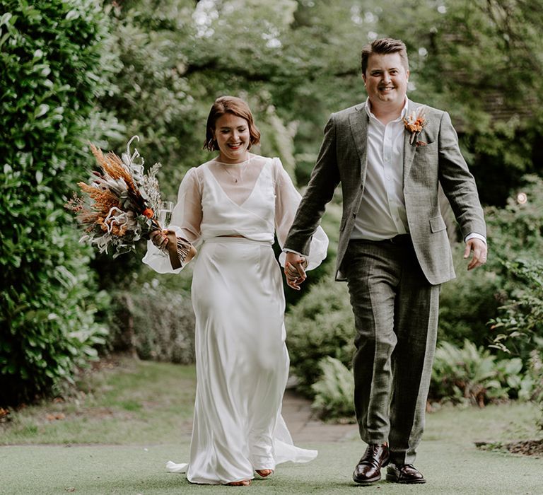 Bride in a satin skirt and camisole with sheer overlay holding hands with her groom in a grey check suit