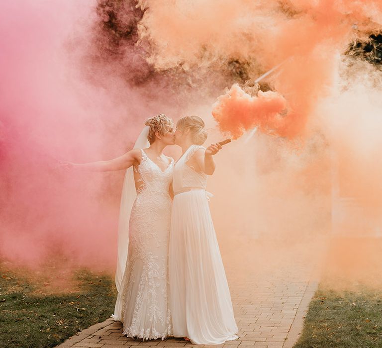 Brides hold pink and orange smoke bombs outdoors 