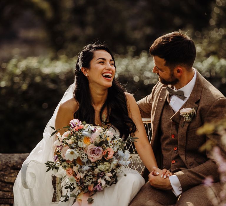 Bride in lace wedding dress and veil holding bright mixed bridal bouquet laughs as she sits with groom in brown tweed three piece suit with floral buttonhole