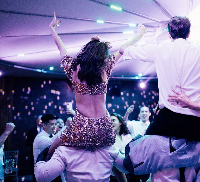 Bride in open back pink glitter wedding dress with bow in hair and groom in white shirt both sit on shoulders of wedding guests on dance floor during evening wedding reception