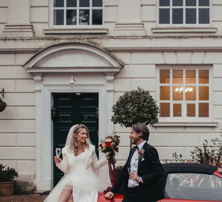 Bride & groom hold champagne glasses in front of red Ferrari at Gosfield Hall 