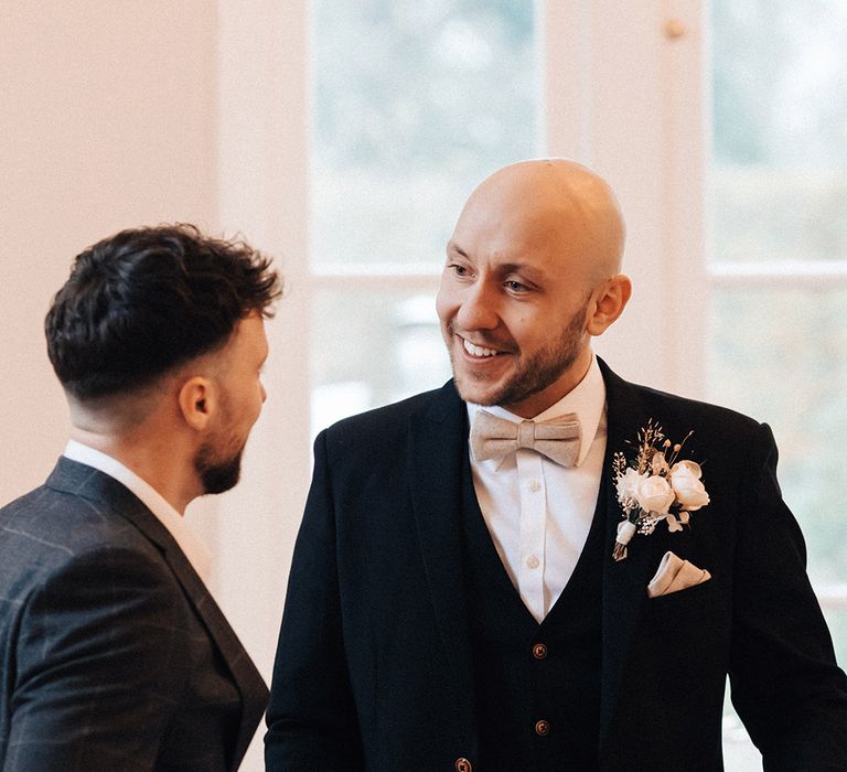 Groom in a black suit and waistcoat with beige bow tie and white flower buttonhole flower 