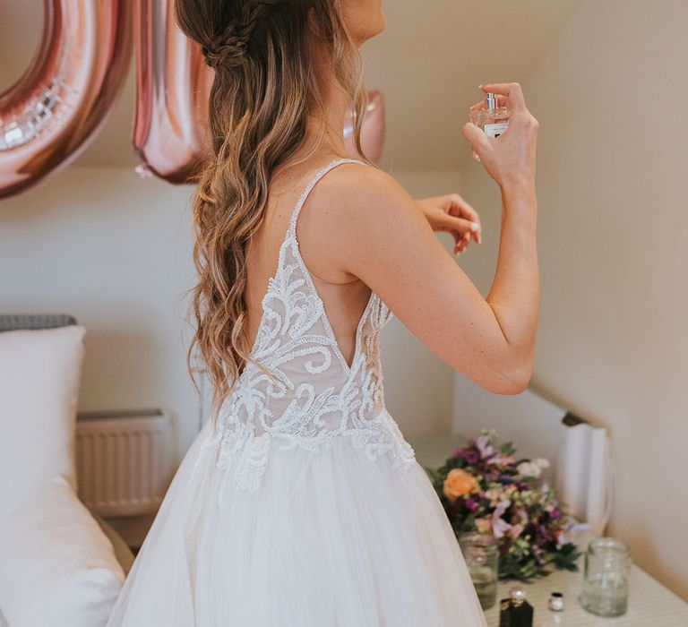 Bride puts on perfume in tulle dress with sparkly bodice and deep v-cut back in front of rose gold balloons 