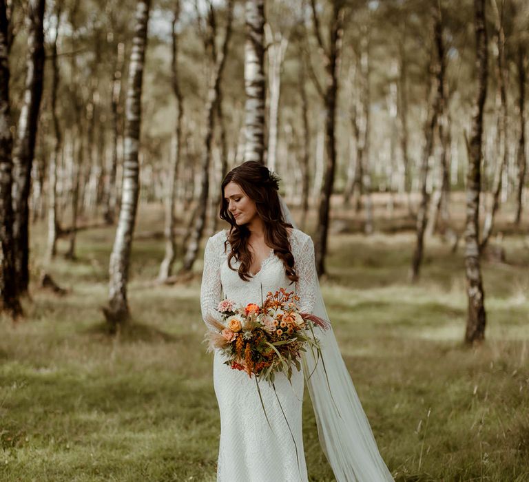 Lace wedding dress with puddle train and embroidered veil holding autumn wedding bouquet in woodland