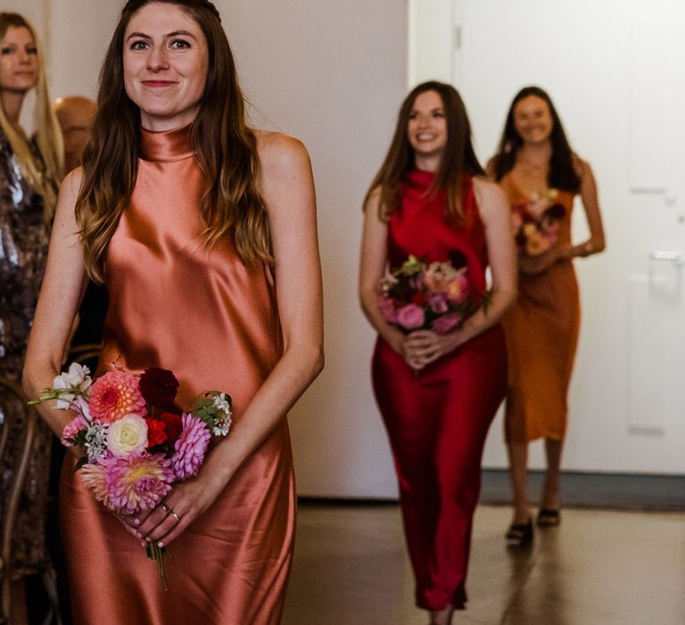 Bridesmaids in orange, red and yellow satin high neck dresses with pink flower bouquet