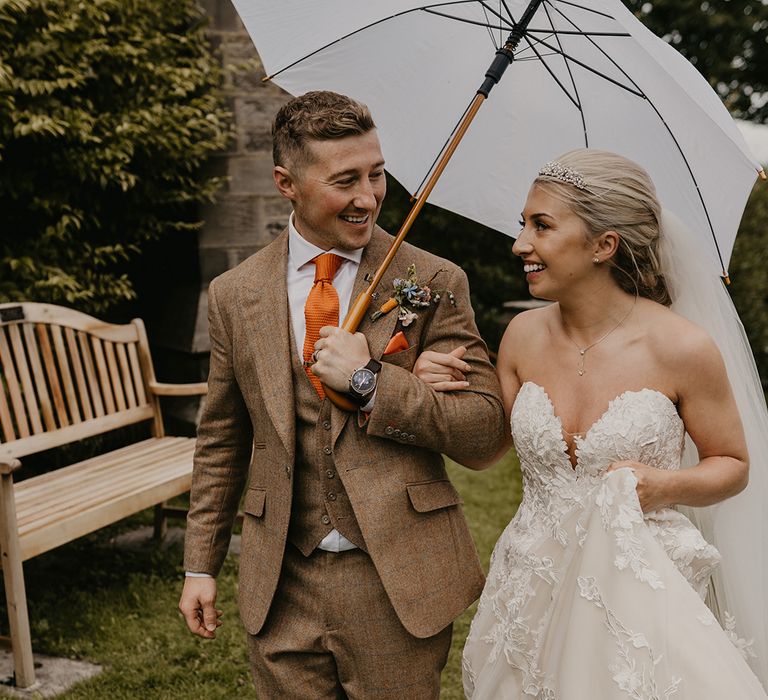 Groom in brown three piece suit with orange tie and handkerchief and colourful boutonnière with white umbrella and bride in sweetheart gown 