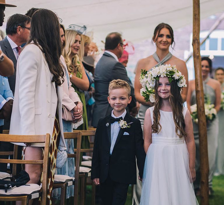 Ring bearer in black tie with white flower buttonhole with flower girl in white dress and large white flower crown walk down the aisle followed by bridesmaids in mint green bridesmaid dresses