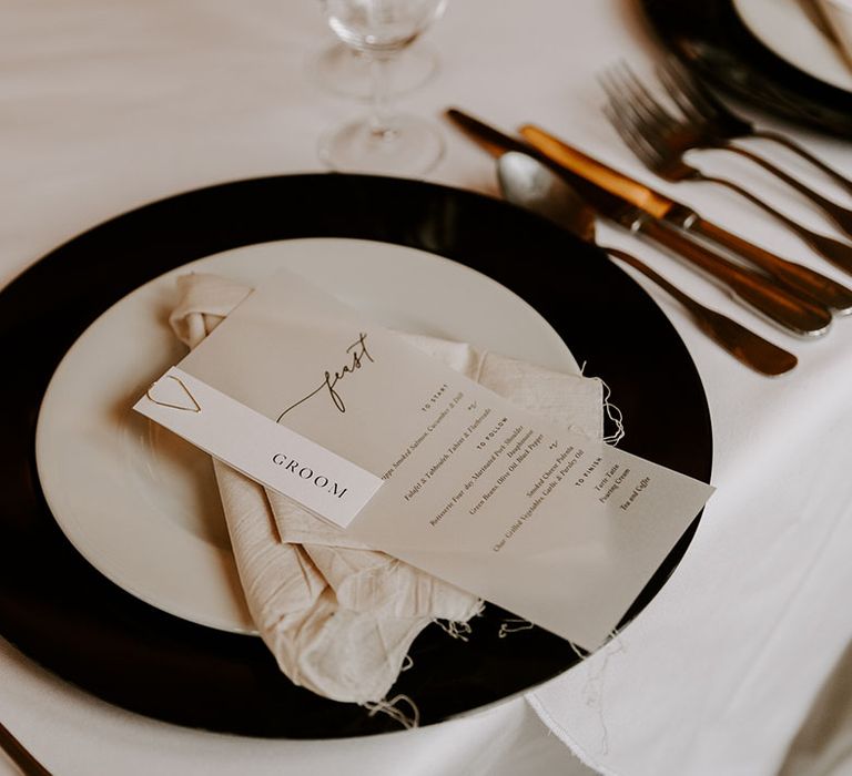 Table setting with black and white plates, gold cutlery, muslin napkin and opaque menu card attached to name place card