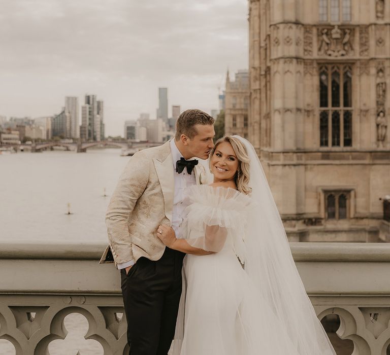 Groom in custom gold patterned tuxedo leans in for a kiss as the bride smiles and looks away in ruffle wedding dress