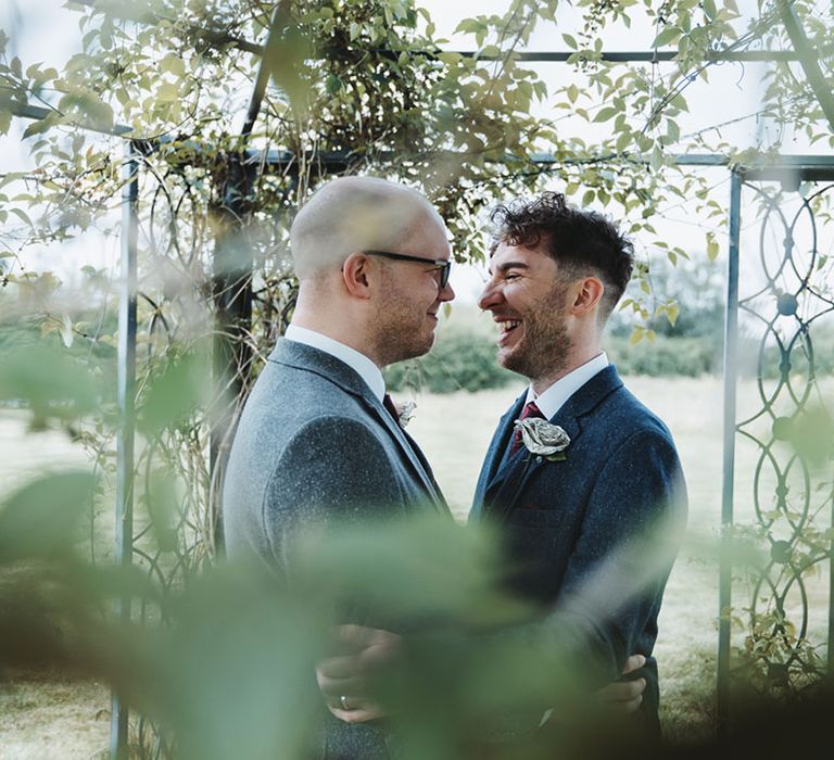 Grooms smile at each other as they stand in the grounds of their wedding venue, Southend Barns