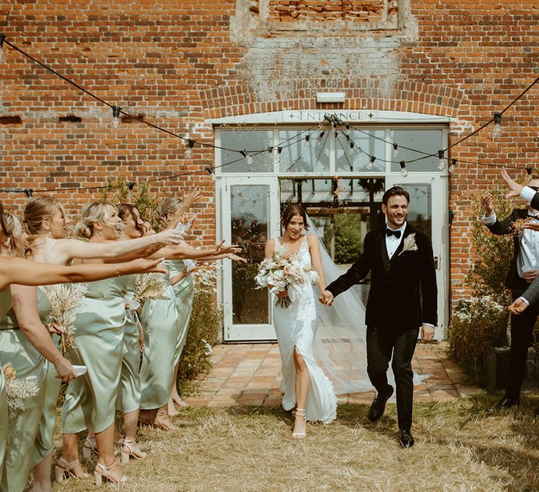 Bride and groom exit from their ceremony to confetti with bridesmaids in sage green dresses and groomsmen in black tie