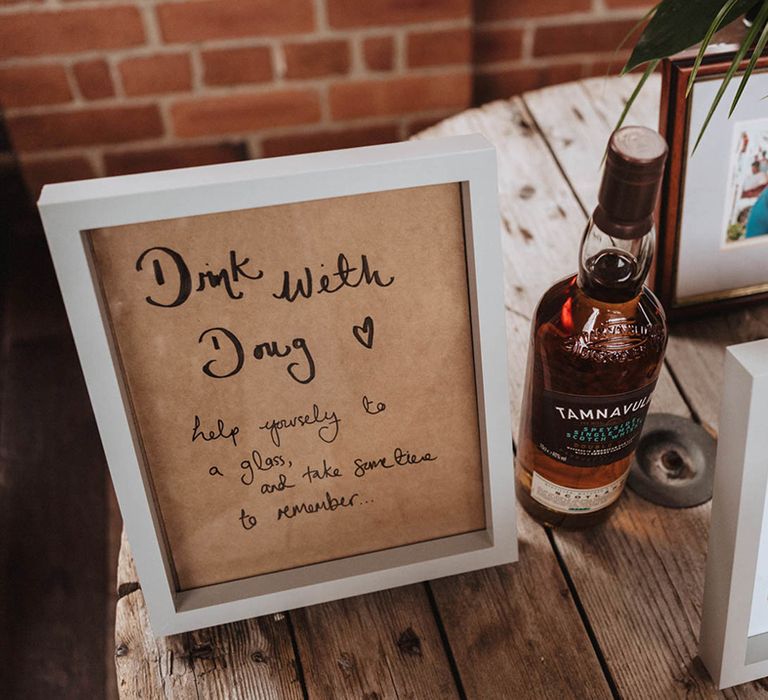 Bottle of spirit next to white photo frame containing sign requesting guests to have a drink for wedding guest who could not be there on the day 