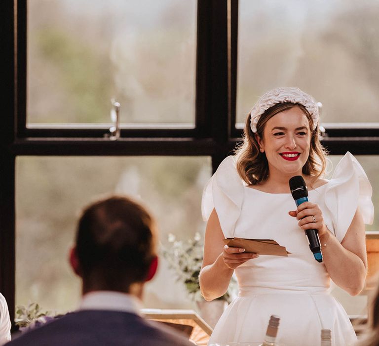 Bride gives a non-traditional bride speech wearing red lipstick and ruffle headband and dress with statement sleeves as groom smiles up at her 