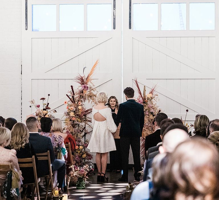 Bride and groom stand at the altar in front of colourful flower columns as guests watch on 