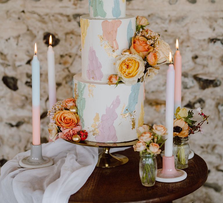 Three-tier white buttercream wedding cake with pastel pink, yellow, green and blue and lilac design surrounded by ombre candles