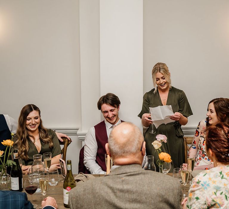 Bridesmaid in green wrap dress gives a speech while everyone listens 