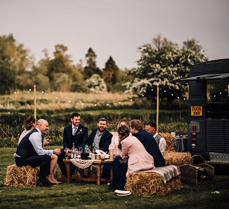 Wedding guests sit on hay bale seats as they talk around a table 