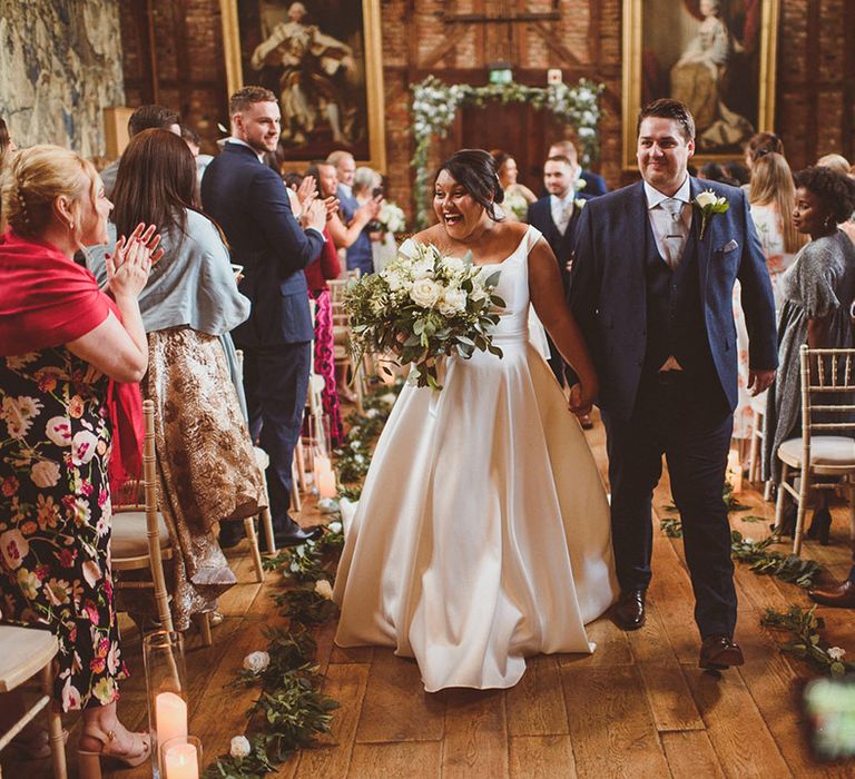 Groom in three piece blue suit and bride in princess skirt wedding dress walk back down the aisle as a married couple