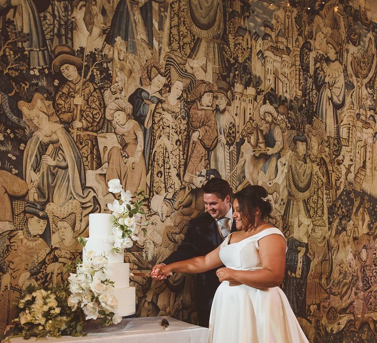 Bride and groom cut their luxe four tier white wedding cake decorated with white flowers and greenery  