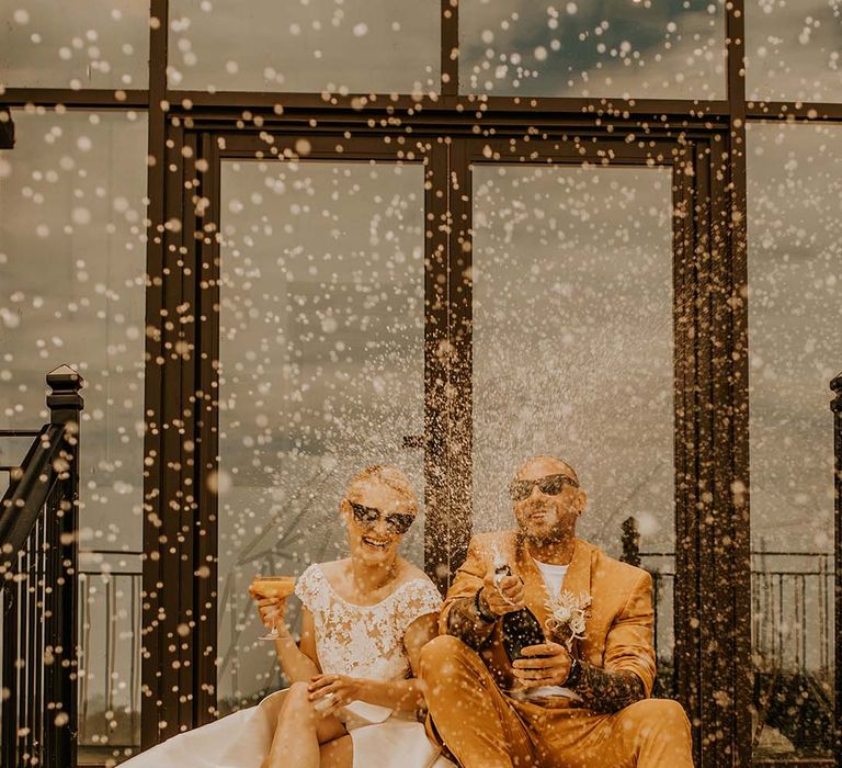 Bride in short sleeved wedding dress with gold heels and groom in mustard suit doing champagne pop