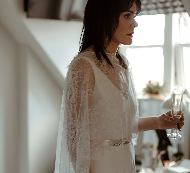 Bride holds glass of champagne and wears dark hair straight alongside lace cape 