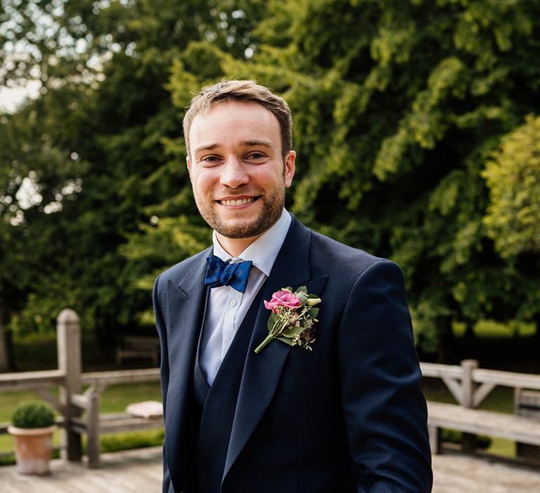 Groom in dark blue suit with blue bow tie with pink rose buttonhole for wedding 