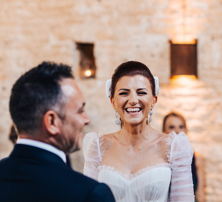 Smiling bride in Emma Beaumont tulle wedding dress with detachable sleeves with ruffles and belt with silver statement earrings with groom at the altar