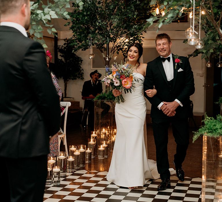 Father of the bride in black tie with red rose buttonhole walks the bride down the aisle at her Larkspur Lodge wedding 