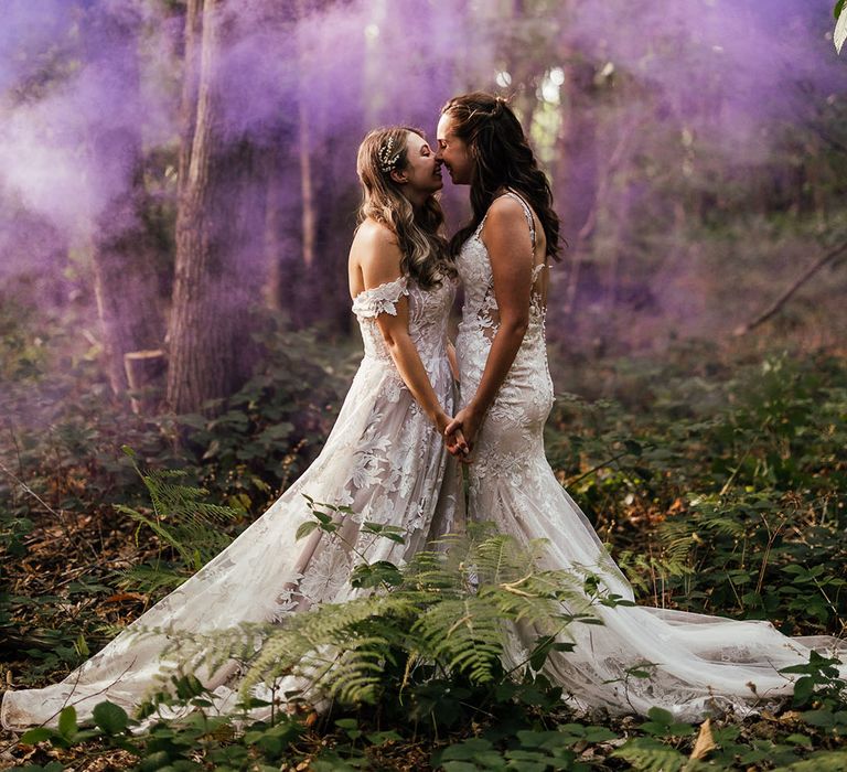 Brides hold hands and lean in for a kiss for stunning purple smoke bomb couple portrait in woodland 